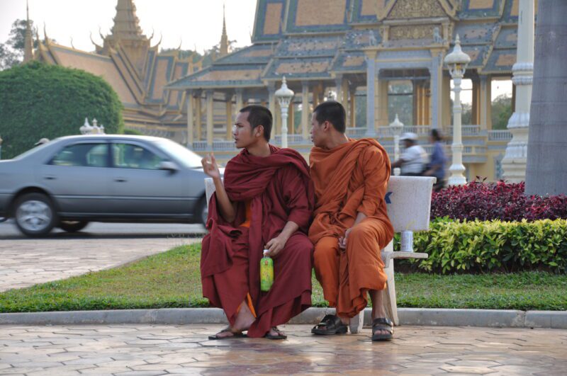 Phnom Penh, Kambodža dvaja mnísi v oranžovom sedia na lavičke, za nimi je auto a vstup do paláca